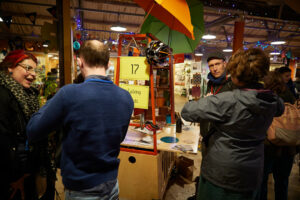 A colour photo of the marketplace at the OAUK 2019 conference. Two conversations are taking place between two groups of people. A table with a colourful flag and promotional material is in the background. 