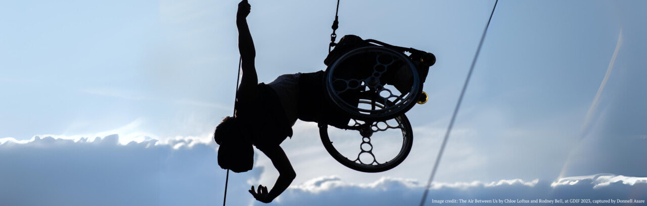 A silhouette of a performer in a wheel chair suspended against a blue sky background. The performer gracefully dances in the air as part of the aerial performance 'The Air Between Us' by Chloe Loftus and Rodney Bell.